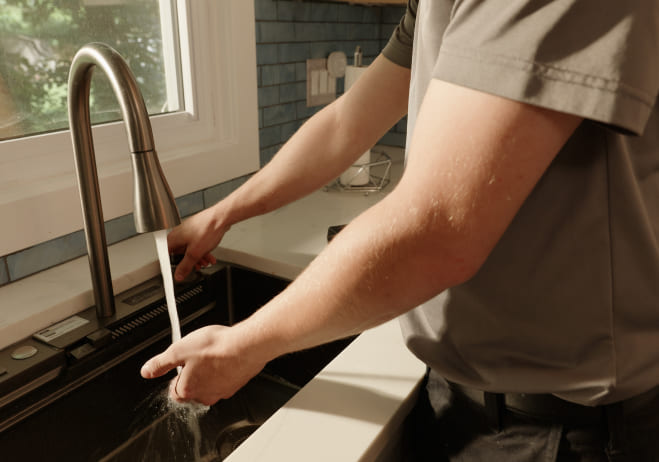 A person in a gray shirt washes hands under a kitchen faucet with running water. Sunlight streams through a window, creating a calm, homely atmosphere.