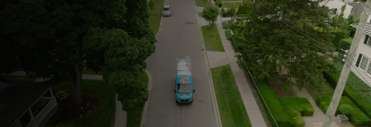 Aerial view of a suburban street with a blue service van driving on it. The street is lined with trees and houses, creating a calm neighborhood scene.