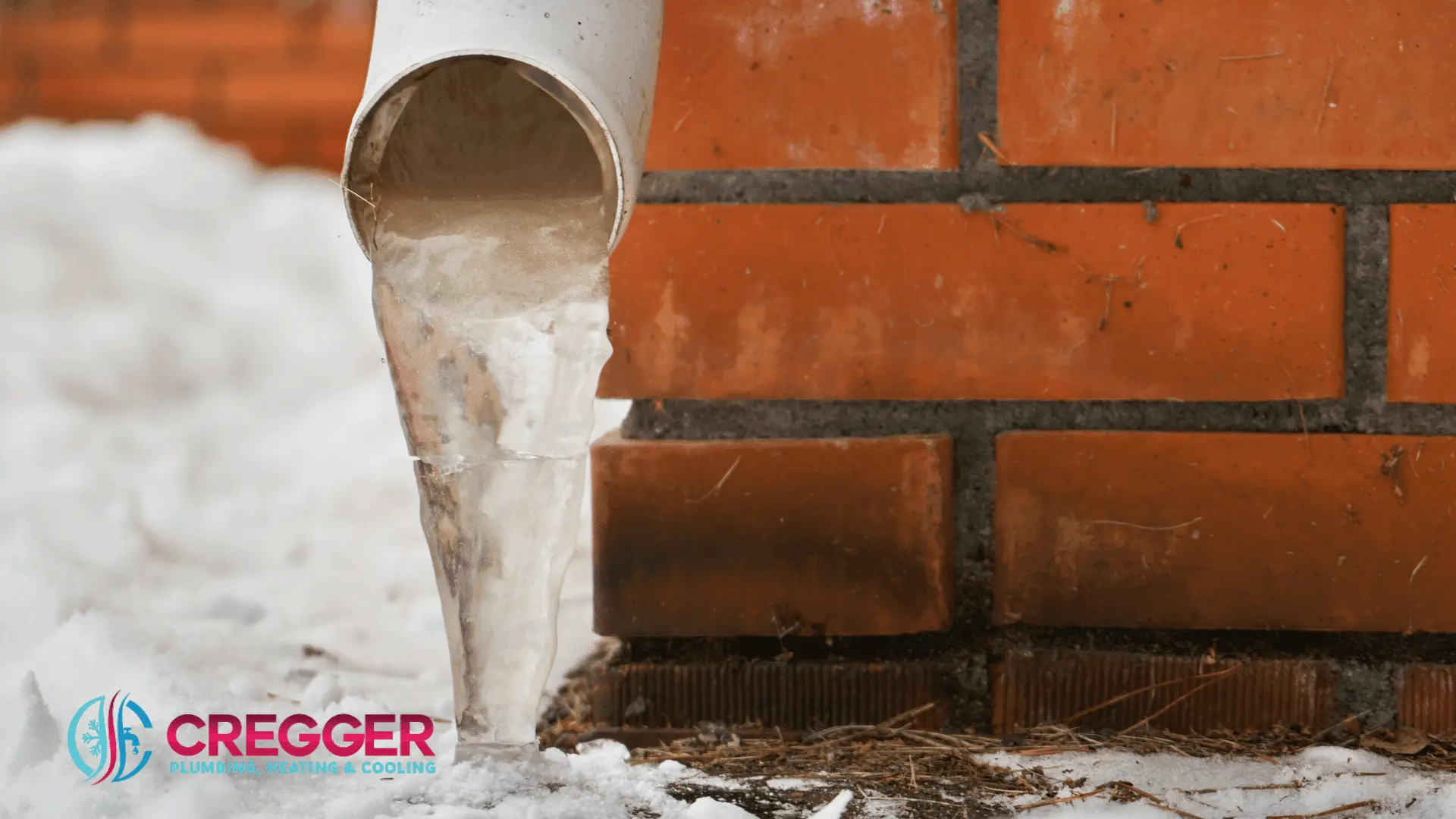 A frozen downspout with icicles extends from a white pipe against a red brick wall. Snow covers the ground, creating a cold, wintry scene.