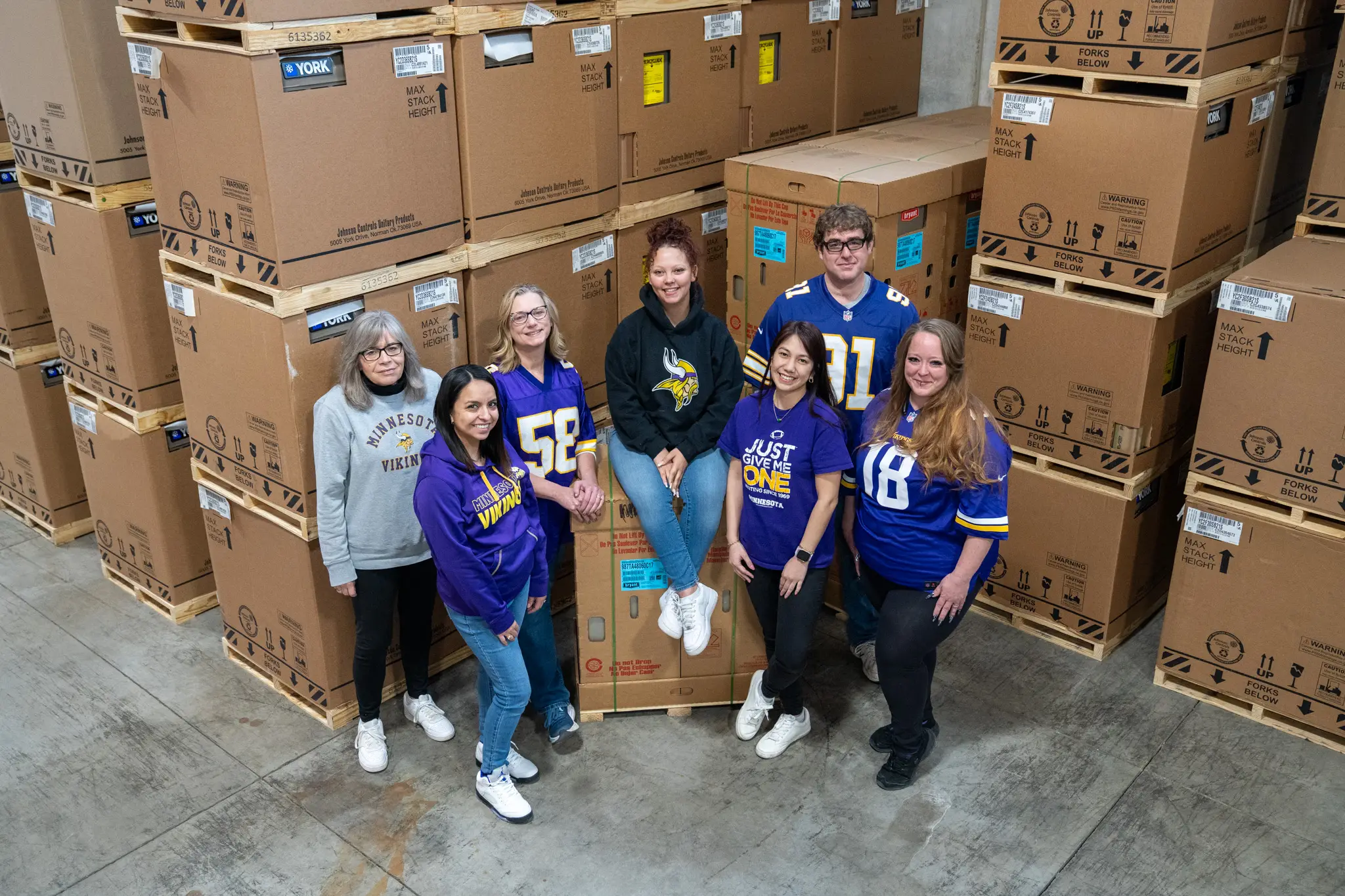 A group of seven people wearing Minnesota Vikings attire stands smiling in a warehouse, surrounded by large stacks of cardboard boxes, conveying a cheerful and team-spirited mood.