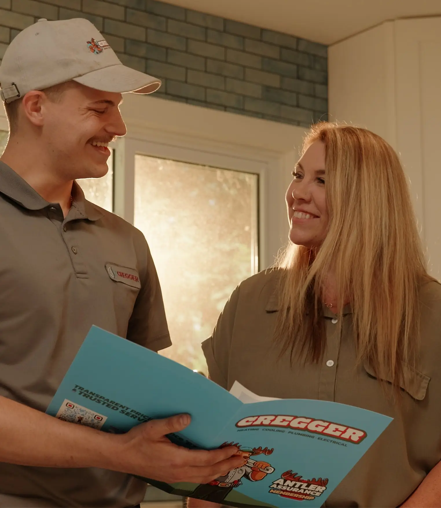 A technician in a uniform and cap smiles at a woman while holding a CREGGER brochure. They stand in a sunlit room, conveying a friendly tone.