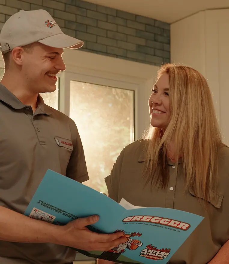 A technician in a uniform and cap smiles at a woman while holding a CREGGER brochure. They stand in a sunlit room, conveying a friendly tone.