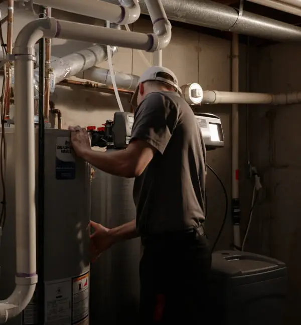 A technician in a dimly lit basement examines a furnace. He wears a cap and uniform, surrounded by pipes and machinery, focused on his work.