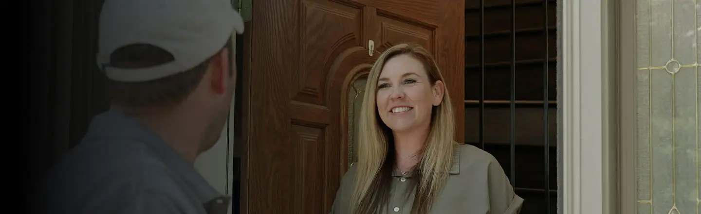 A woman with long blonde hair smiles warmly at a person wearing a cap outside her door. The brown wooden door is partially open, creating a welcoming tone.