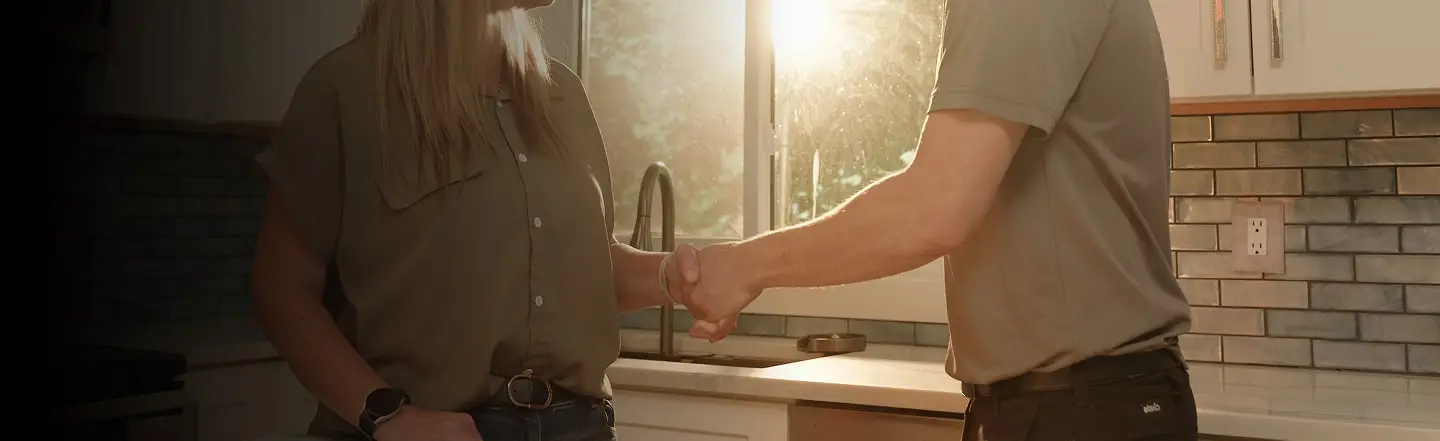 Two people shake hands in a sunlit kitchen, conveying a sense of agreement or partnership. Warm light filters through a window, creating a cozy atmosphere.
