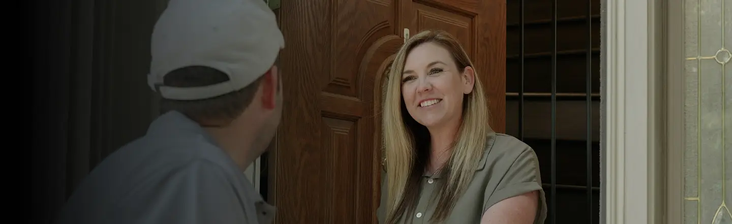 A woman with long hair smiles warmly at a man in a cap standing at a wooden door. The scene conveys a friendly and welcoming atmosphere.